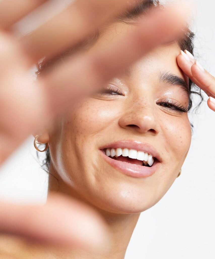Jeune femme brune aux cheveux bouclés, radieuse, main devant la moitié de l’objectif – Sahiya Method, confiance et bien-être.