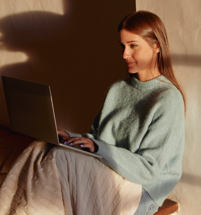 Jeune femme châtain clair assise, couverte d’un plaid, travaillant sur son laptop – Sahiya Method, lifestyle et bien-être équilibré.